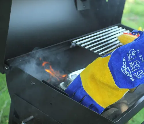 Hand in a RAPICCA blue and tan welding glove adjusting a grill with live coals.
