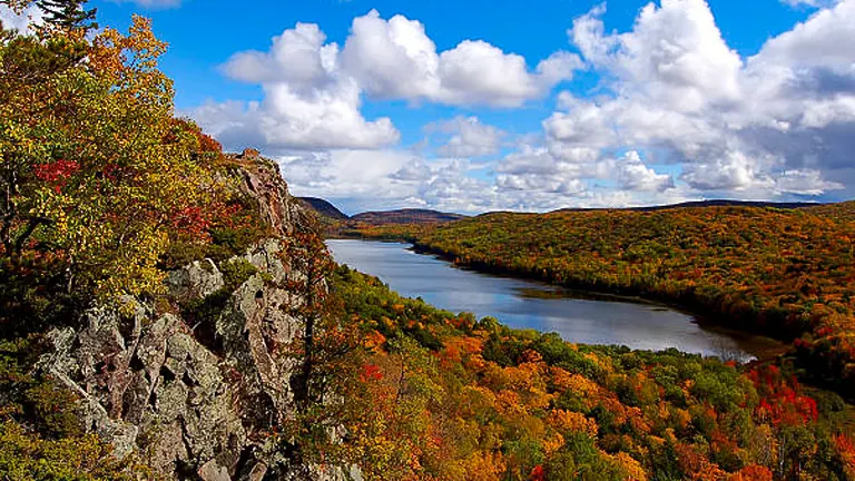 A vibrant autumn landscape with a river winding through colorful forested hills under a bright blue sky with fluffy clouds.