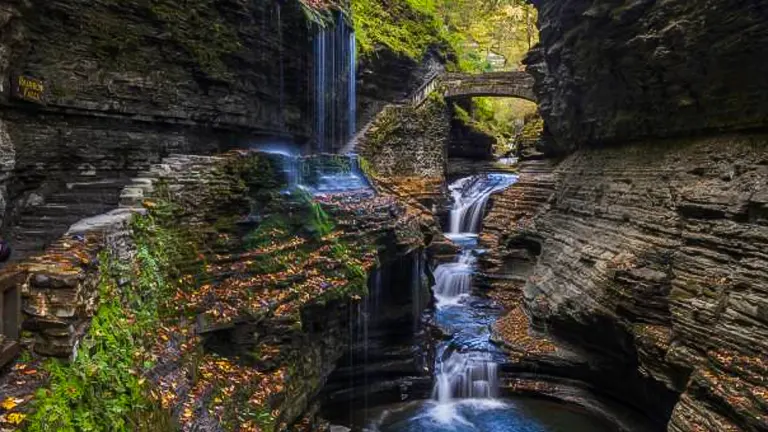 Waterfalls cascade down a rocky gorge with autumn leaves scattered around and a stone bridge arching overhead.