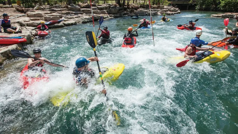 A group of kayakers navigating through turbulent white water rapids, with some paddlers actively maneuvering and others watching from the riverbank, surrounded by lush greenery.