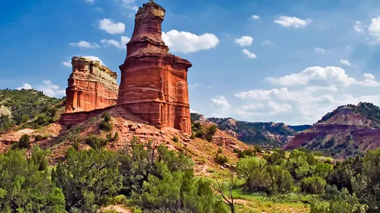 A striking red rock formation stands prominently amid green shrubbery under a partly cloudy sky.
