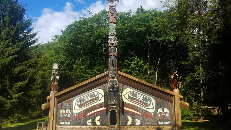A traditional Alaskan clan house with a richly decorated façade and totem poles on either side, set against a lush forest backdrop under a clear sky.