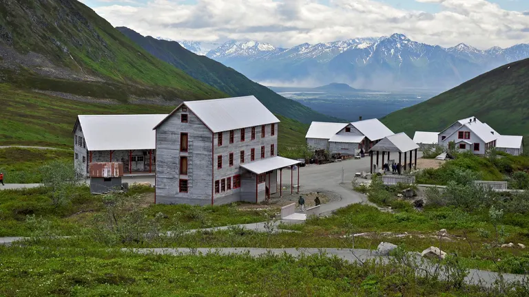 Historic mining buildings with white walls and red windows at Independence Mine State Historical Park in Hatcher Pass, Alaska, set against a backdrop of green mountains and cloudy skies.