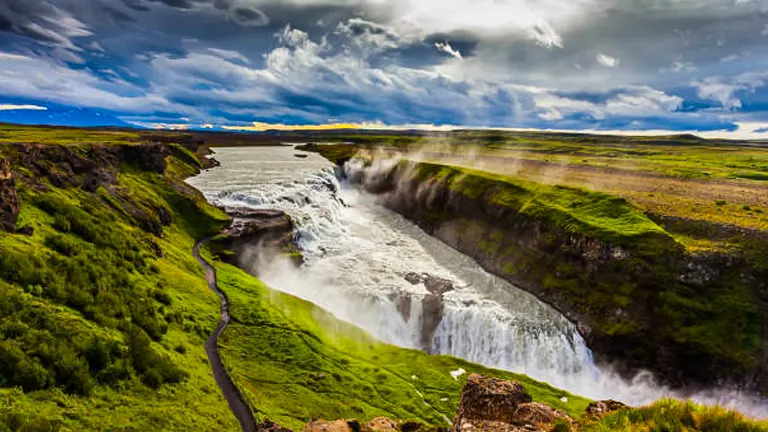 Dynamic view of Gullfoss Waterfall in Iceland, with the Hvítá River flowing powerfully through green terrains under a dramatic sky with sun rays piercing through clouds.
