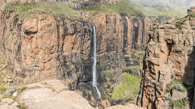 View of the slender Tugela Falls gracefully descending the rugged cliffs of the Drakensberg Mountains, under a clear sky, with trails visible in the surrounding landscape.