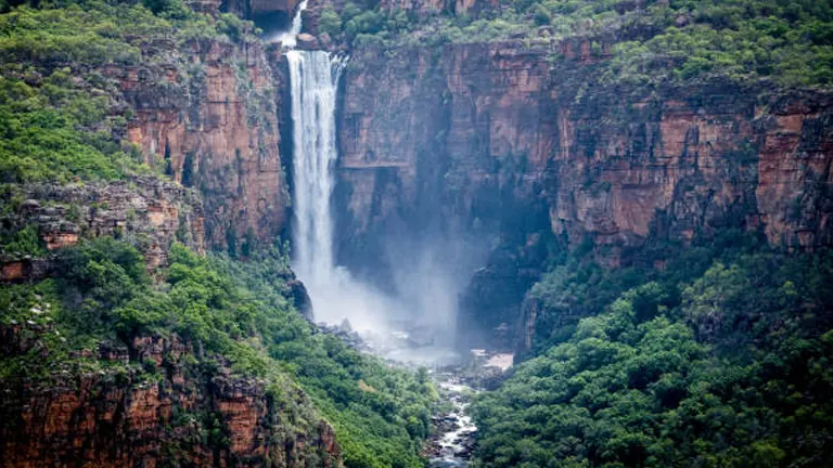 Twin Falls in Kakadu National Park, Australia, with water plunging down high red cliffs into a mist-filled gorge surrounded by lush greenery.