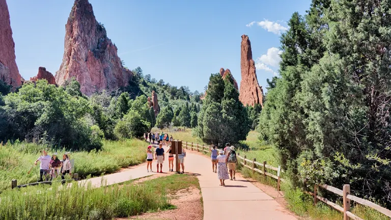Visitors walking on a paved trail in a park with towering red rock formations on either side, lush green trees lining the path under a clear blue sky.