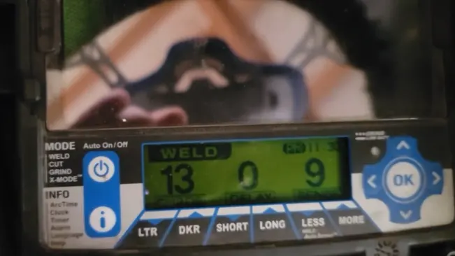 Close-up of the digital control interface on a Miller Digital Infinity welding helmet, showing shade level and modes with reflection of a welder in the screen."