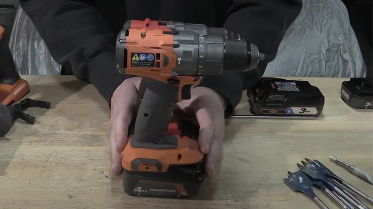 Hands securing a battery into a RIDGID cordless hammer drill on a workbench with drill bits and a spare battery in view.