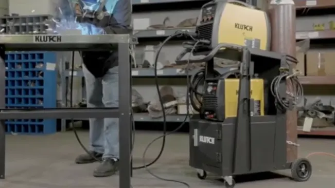 Alt Text: "Klutch 2-Tier Welding Cart with equipment and accessories in a workshop, with a welder working in the background.