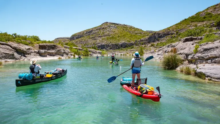 Kayakers on a clear, calm river gently flowing through a rugged limestone canyon under a bright blue sky, illustrating a peaceful day of water exploration in a scenic outdoor setting.