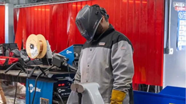 Welder in a Lincoln Electric XVI Series Industrial Jacket and welding helmet, working in a shop environment.






