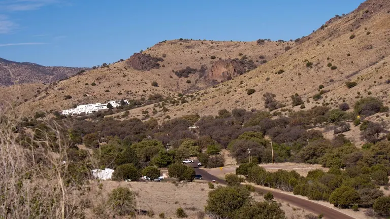 A landscape showing a winding road leading to a white building nestled in the foothills of arid, tree-covered mountains under a clear blue sky.
