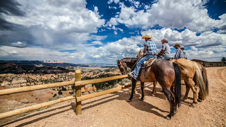 Two riders on horseback pause to enjoy a panoramic view of a rugged canyon under a vast sky with dramatic clouds.