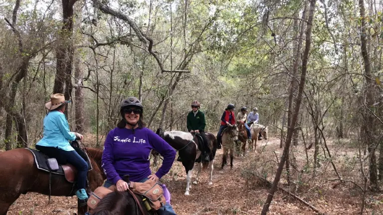 A group of smiling riders on horseback winding through a dense, leafy forest trail, with the lead rider looking towards the camera.

