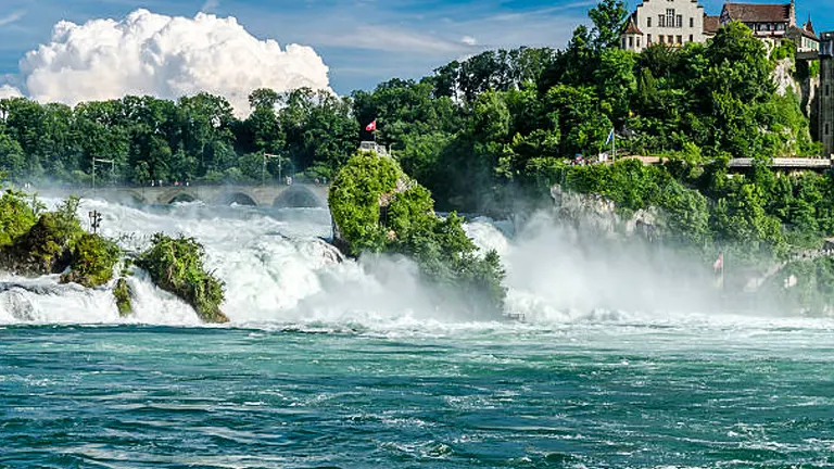 The Rhine Falls in full flow, with mist hovering above the emerald waters and Schloss Laufen on the clifftop, set against a backdrop of a clear blue sky with fluffy clouds.