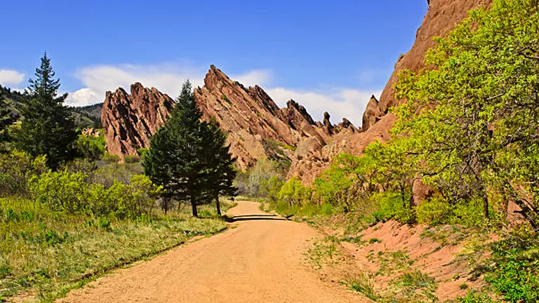 A dirt path leading through a scenic landscape with distinctive red rock formations on one side and lush greenery on the other, under a clear blue sky.