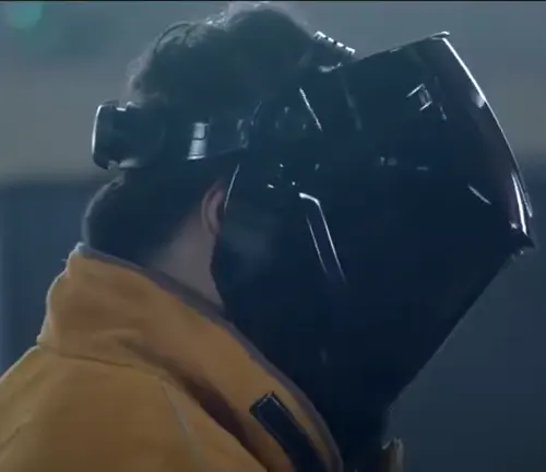 Side view of a welder wearing a black welding helmet and a yellow jacket.






