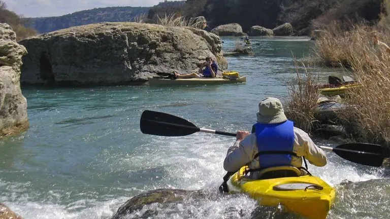 A kayaker in a yellow kayak navigates through small rapids in a river with large rocks and calm pools, surrounded by a rugged landscape under a partly cloudy sky.