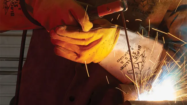 Close-up of hands in yellow welding gloves holding a rod and creating sparks while wearing a brown leather welding jacket.