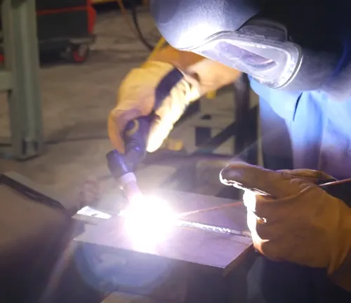 Welder using a TIG torch to weld metal, with bright light from the arc, wearing protective gloves and possibly using a 3M Speedglas G5-01 helmet.




