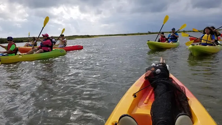 A first-person perspective of a kayaker's legs in a red kayak, leading a group of kayakers with yellow paddles across a choppy coastal waterway under an overcast sky.