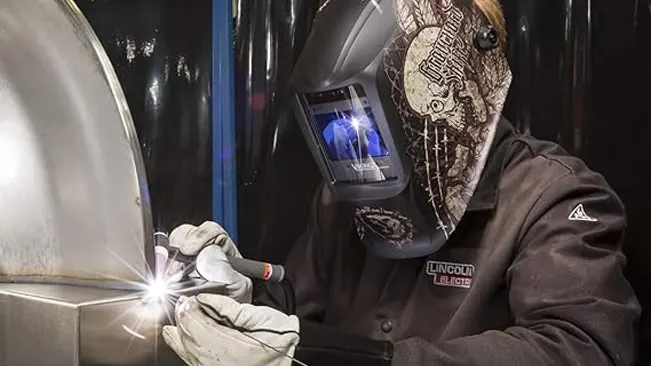 Welder in a camouflage-patterned helmet and Lincoln Electric K2985 jacket performing an arc welding process.