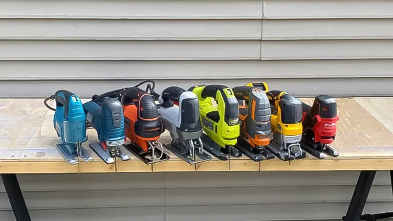 A lineup of eight different jigsaw tools in various colors and brands displayed on a wooden plank against a house siding