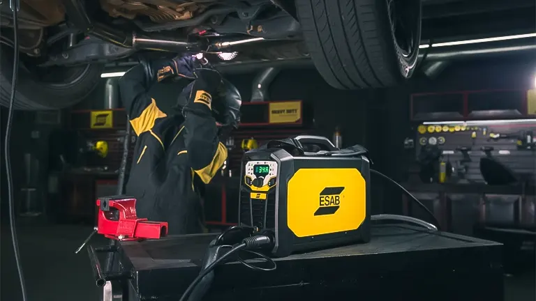A welder in protective gear using an ESAB stick welder beneath a vehicle in a workshop