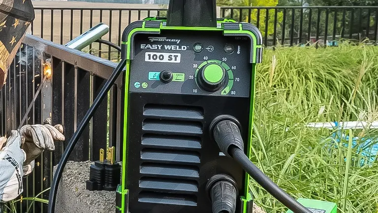 A close-up of a Forney Easy Weld 100 ST stick welder with its control panel visible, set up for welding outdoors