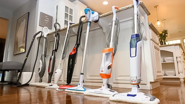 A lineup of five floor cleaning machines with varying designs on a hardwood floor in a residential kitchen