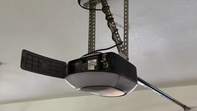 Underneath view of a mounted garage door opener with a manual release handle and built-in light.