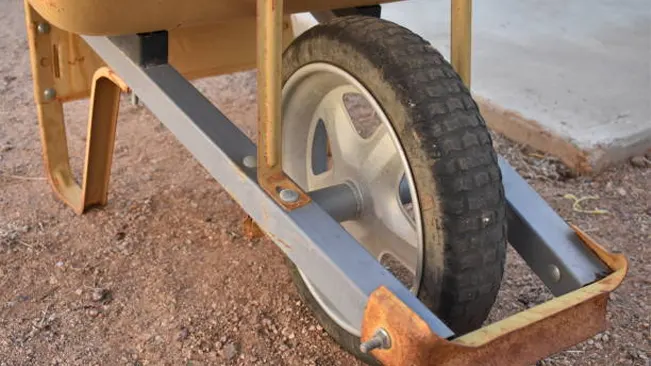 Close-up of a wheelbarrow's single wheel with a rusted metal bracket and pneumatic tire on a dusty ground.