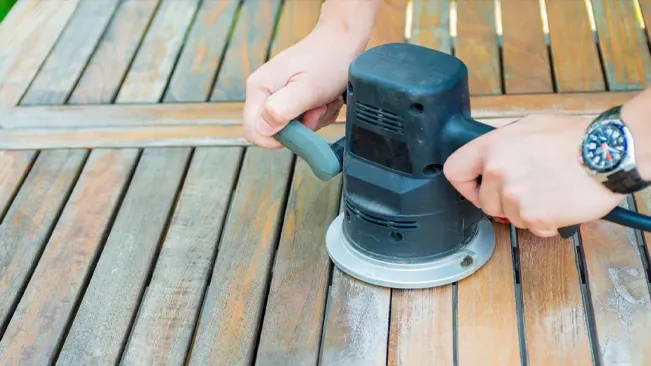 Hands using an orbital sander on a wood deck for post-washing treatment.