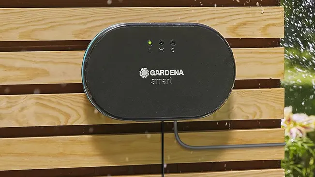 Close-up of a wall-mounted GARDENA smart irrigation control device with indicator lights on, set against a wooden slat background