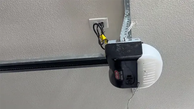 Close-up of a black Genie Chain Drive 500 garage door opener with a white cover, mounted on a metal bracket next to an electrical outlet