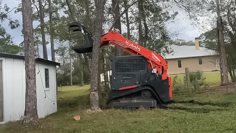 Red Kubota skid steer loader near a white shed in a wooded area