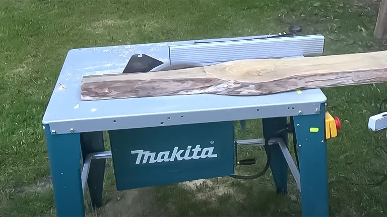 Outdoor setup of a Makita 2712 table saw cutting through a plank of wood, with a visible blue base and tabletop, and a rip fence to the side