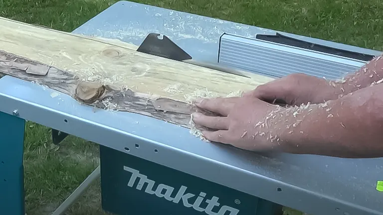 Close-up of hands guiding a wood plank through a Makita 2712 table saw, with sawdust scattered on the table