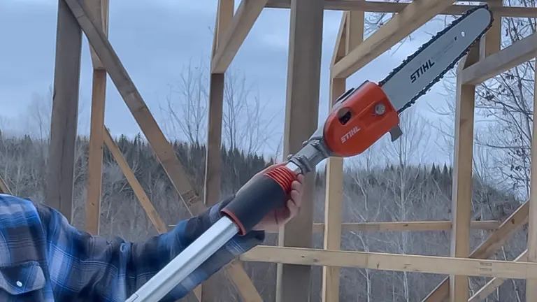 Person holding a STIHL HT 135 pole saw with a wooden frame structure in the background