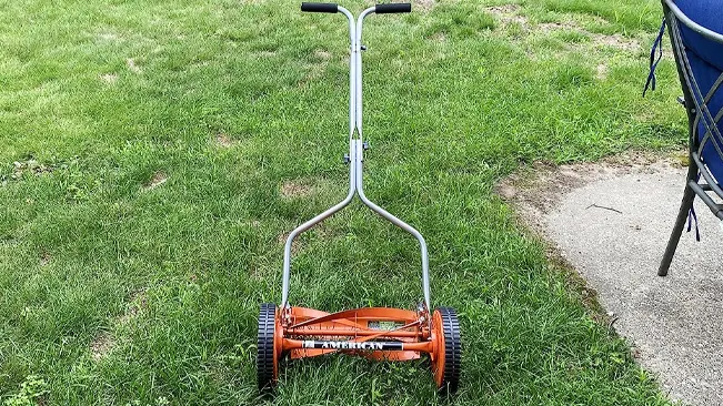 manual push lawn mower with an orange base and silver handlebars, positioned on a grassy area with unevenly cut grass