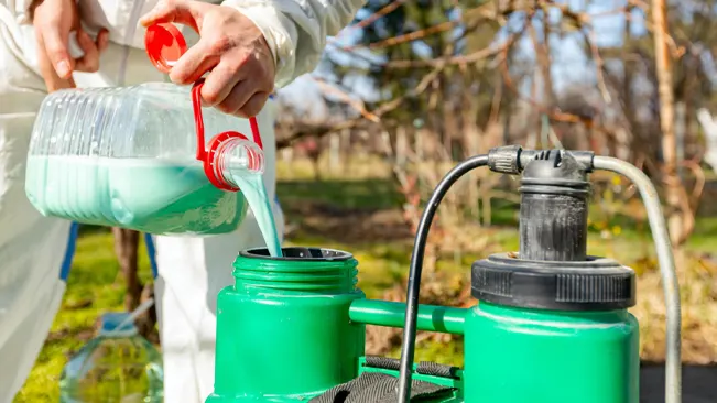 person, visible from the waist down, pouring a light blue liquid from a clear plastic bottle into a green sprayer tank