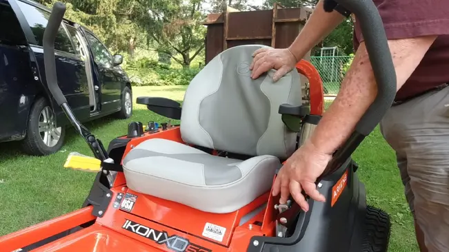 A person adjusting the seat of an orange IKON-XD lawnmower parked on a grassy area