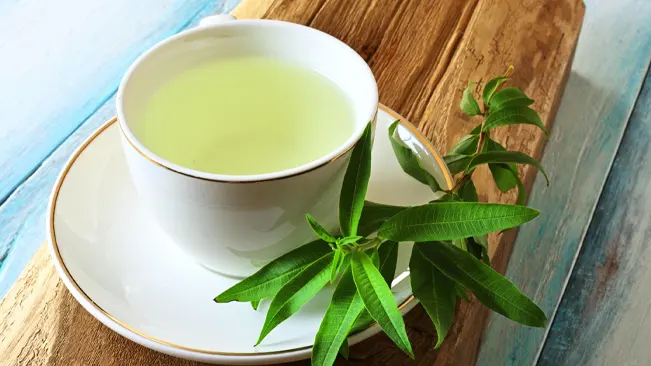A cup of lemon verbena tea beside fresh verbena leaves on a rustic wooden table, illustrating the plant's herbal benefits.