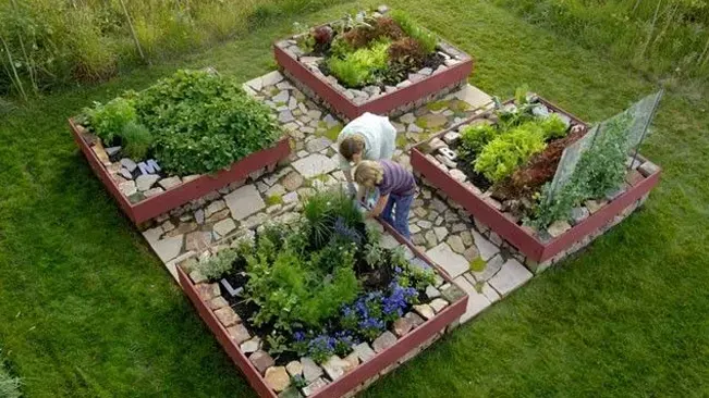An overhead view of a four-square garden layout with raised beds bordered by red bricks, featuring a variety of plants, and a person tending to the central bed.