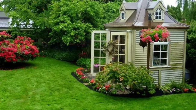 Victorian-style garden shed surrounded by lush flowering plants and a well-manicured lawn.