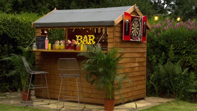 Backyard wooden bar shed with stools and neon 'BAR' sign, for outdoor entertainment.