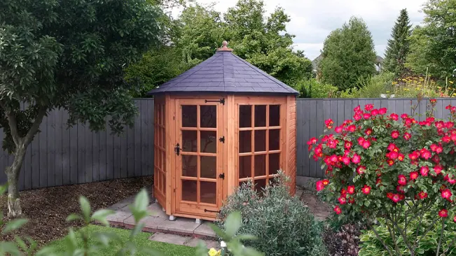 Octagonal wooden shed with round roof in a garden with blooming roses.