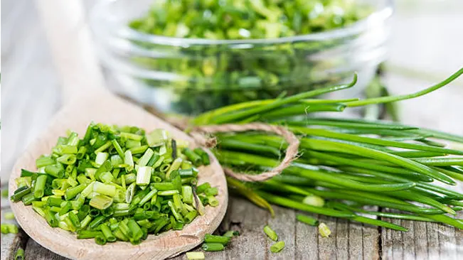 Freshly chopped chives on a wooden spoon with whole chives and a glass bowl in the background.
