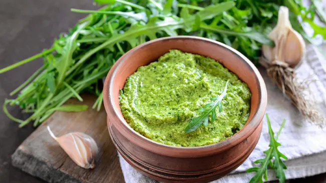Arugula pesto in a terracotta bowl with fresh arugula leaves and garlic in the background, highlighting its culinary uses.